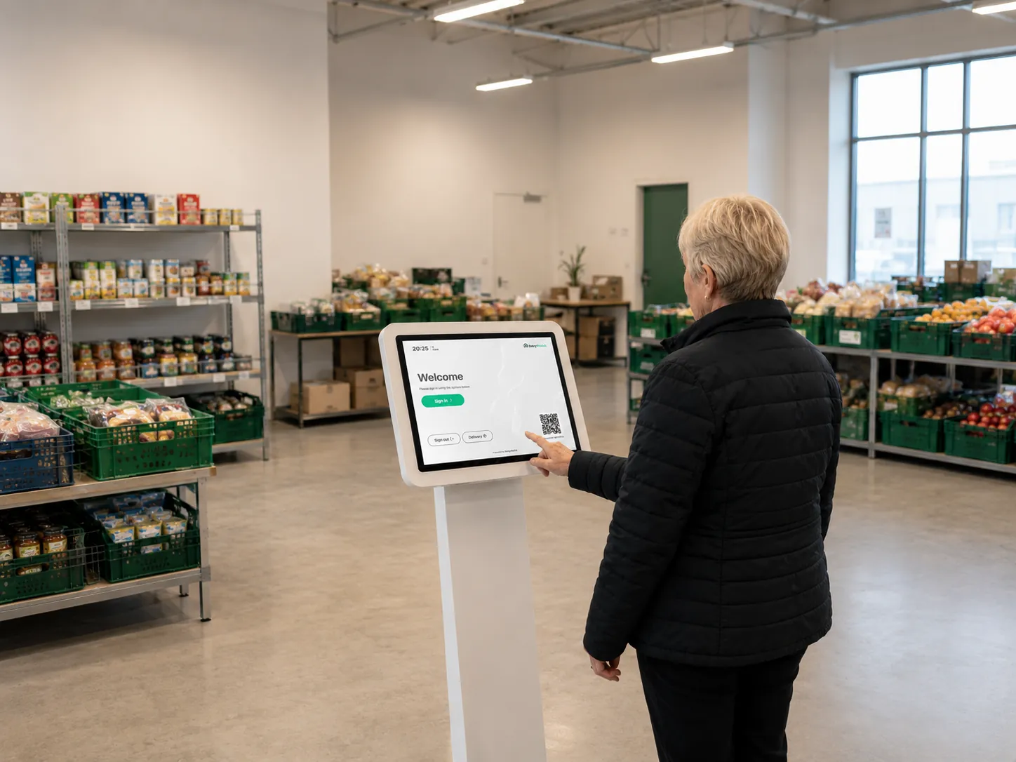 A volunteer in a warm jacket signing in at a floor-standing EntryWatch kiosk in a food distribution centre, shelves of tinned food and crates of fresh produce behind her