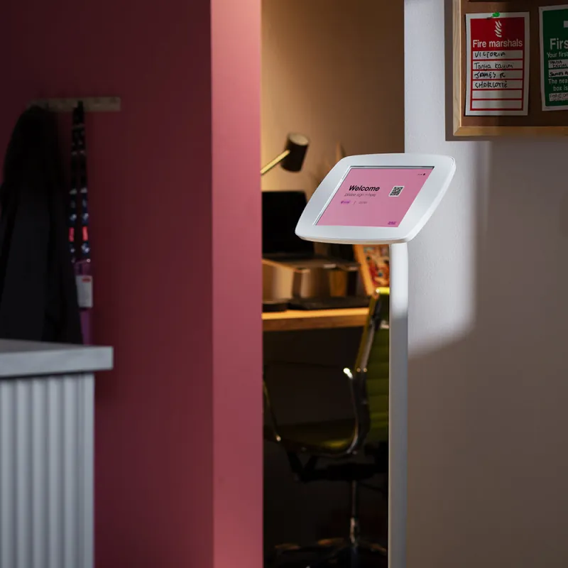 A white Bouncepad floor stand in an office reception, its screen reading “Welcome — please sign in here” beside a QR code